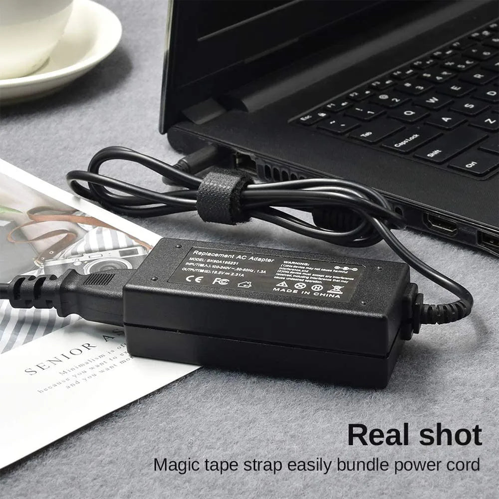 A black universal laptop power adapter with a magic tape strap is shown next to an open laptop.