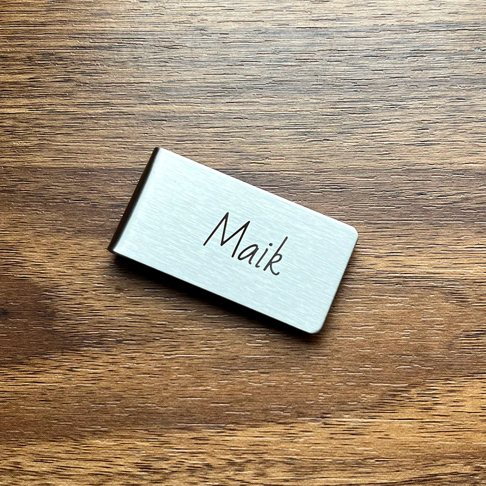 A silver engraved money clip wallet with the name Maik resting on a wooden surface.