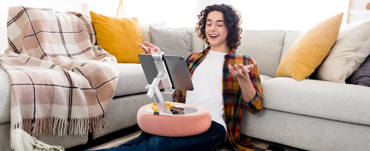 A young man is smiling while holding an adjustable pillow tablet and Kindle stand with a soft plush fabric on a sofa.