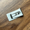 A silver personalized engraved money clip wallet lying on a dark wooden surface.