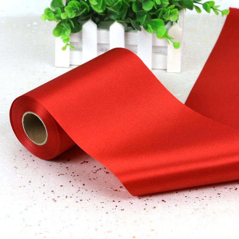 A roll of vibrant red satin ribbon lying on a white surface with greenery in the background.
