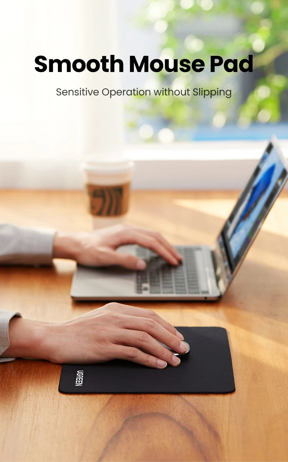 A hand is using a black non-slip rubber mouse pad on a wooden desk in front of an open laptop.