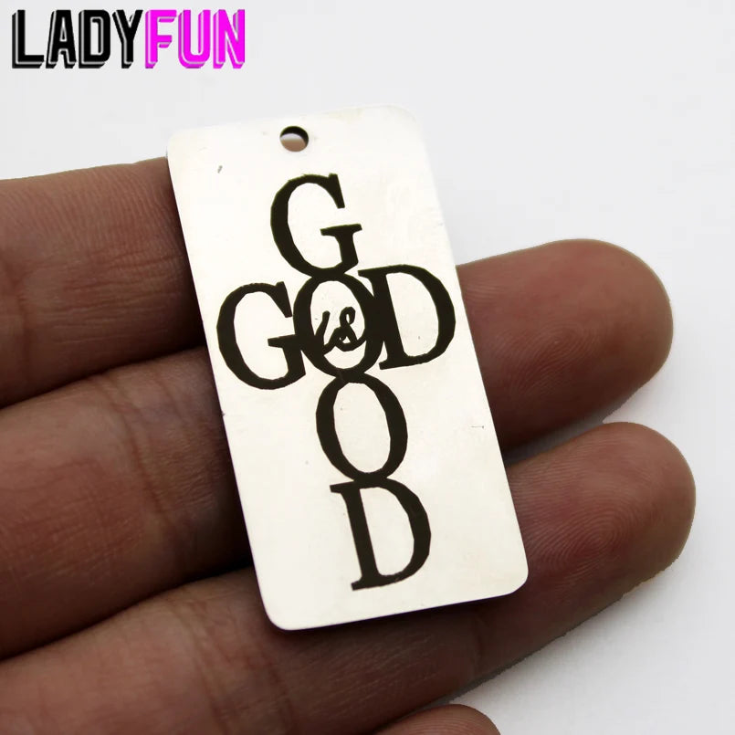 A white rectangle charm pendant engraved with the text God Is Good is held in a hand.