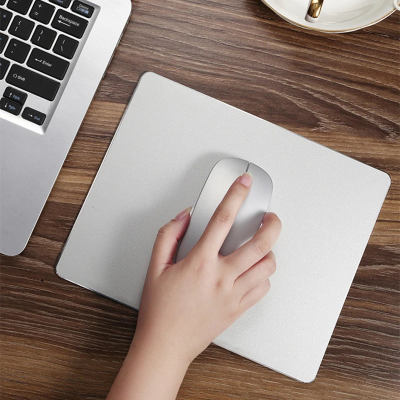 A hand is placing a silver mouse pad on a wooden desk next to a laptop keyboard.