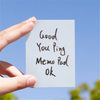 A hand holds a white memo pad with the handwritten text Good You Ping Memo Pad Ok against a blue sky background.