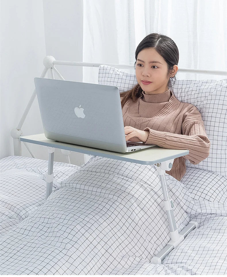 A woman is using a portable adjustable laptop desk stand to position a silver laptop on her lap while sitting on a bed.