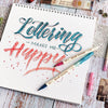 A set of three retro twin markers for watercolor art is displayed on a notebook with lettering and various art supplies.