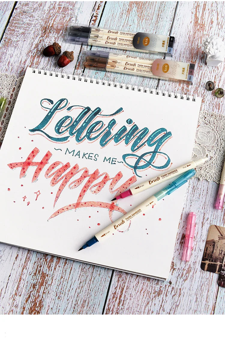 A set of three retro twin markers for watercolor art is displayed on a notebook with lettering and various art supplies.