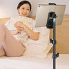 A woman is reading a book while resting on a bed, using an adjustable phone and tablet tripod mount.
