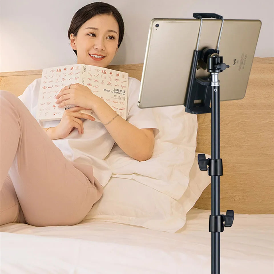 A woman is reading a book while resting on a bed, using an adjustable phone and tablet tripod mount.