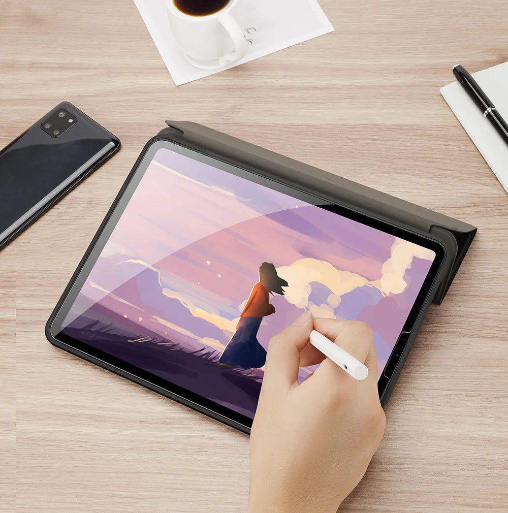 A hand is writing on a tablet displaying a sunset landscape, next to a smartphone and a pen.