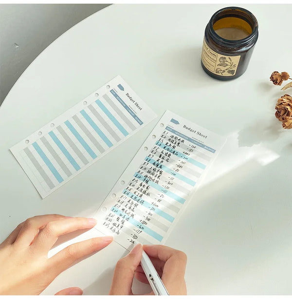 A hand holds a budget sheet with various colored cards and a pen on a white table next to a jar.