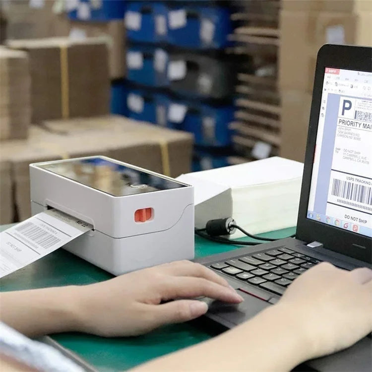 A person is printing thermal shipping labels using a handheld device next to a laptop in a warehouse setting.