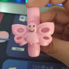 A hand holds a pink silicone pencil grip shaped like a smiling butterfly for handwriting aid.