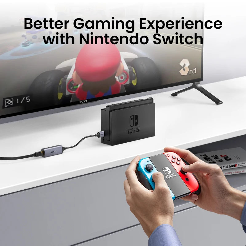 A hand holds a Nintendo Switch console and an adapter plugged into a computer setup for gaming.