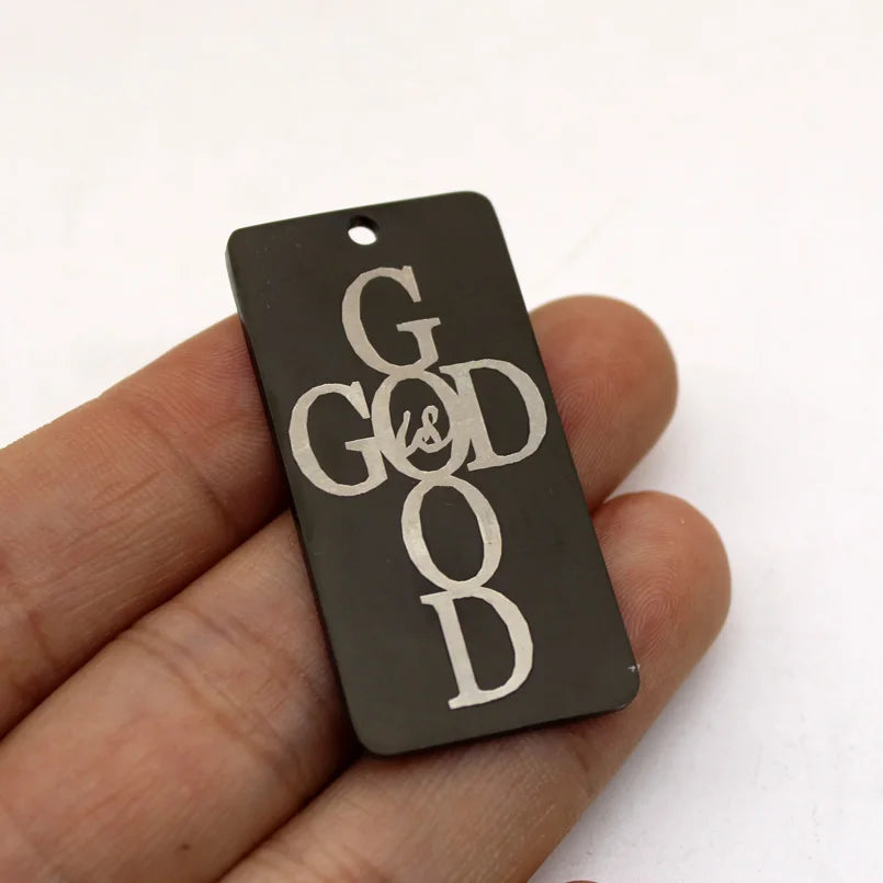 A dark rectangle stainless steel charm pendant engraved with the words God Is Good held in a hand.