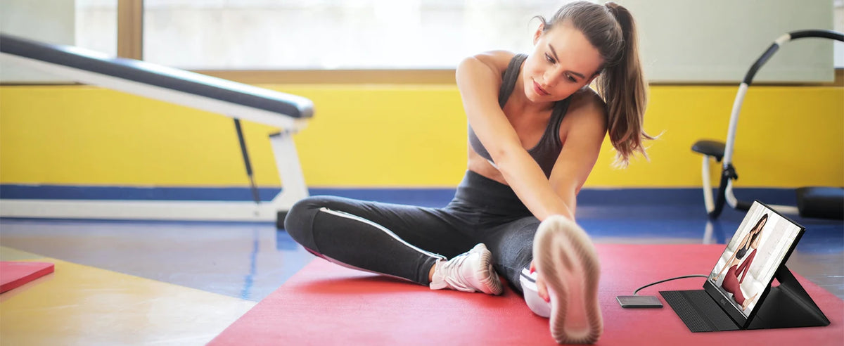 A woman is using a red yoga mat and a portable laptop monitor in a brightly lit gym setting.