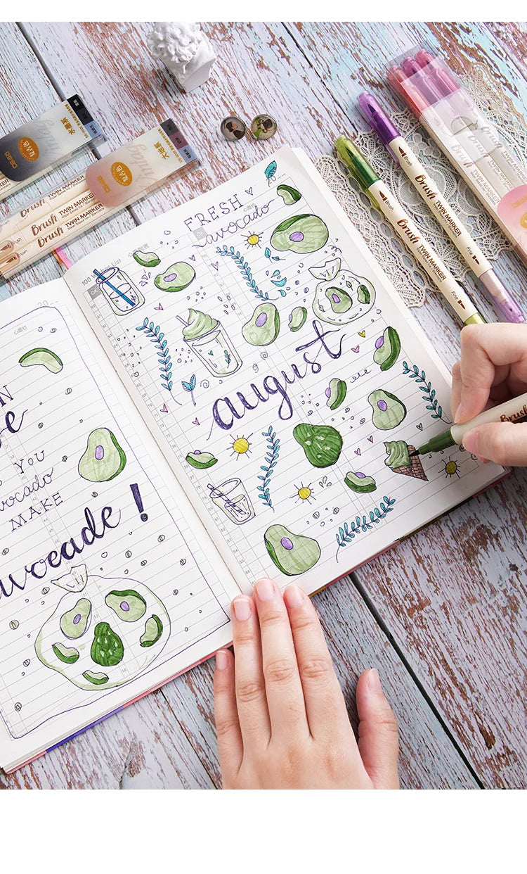 A hand writes 'FRESH AUGUST' in watercolor style on a notebook next to a set of three retro twin markers.