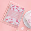 A pink clear plastic planner with a floral pattern and a pink cover, featuring a 100 Envelope Savings Challenge.
