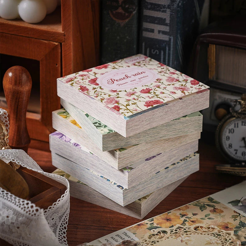 A stack of vintage floral square scrapbook paper sheets with pink and green patterns resting on a wooden surface.
