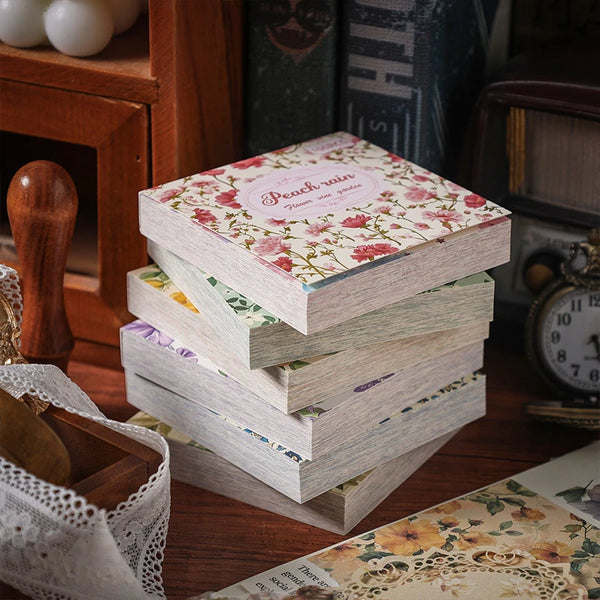 Stapel alter Blumen-Quadrat-Scrapbook-Papierblätter mit rosa und grünen Mustern auf einer Holzoberfläche.
