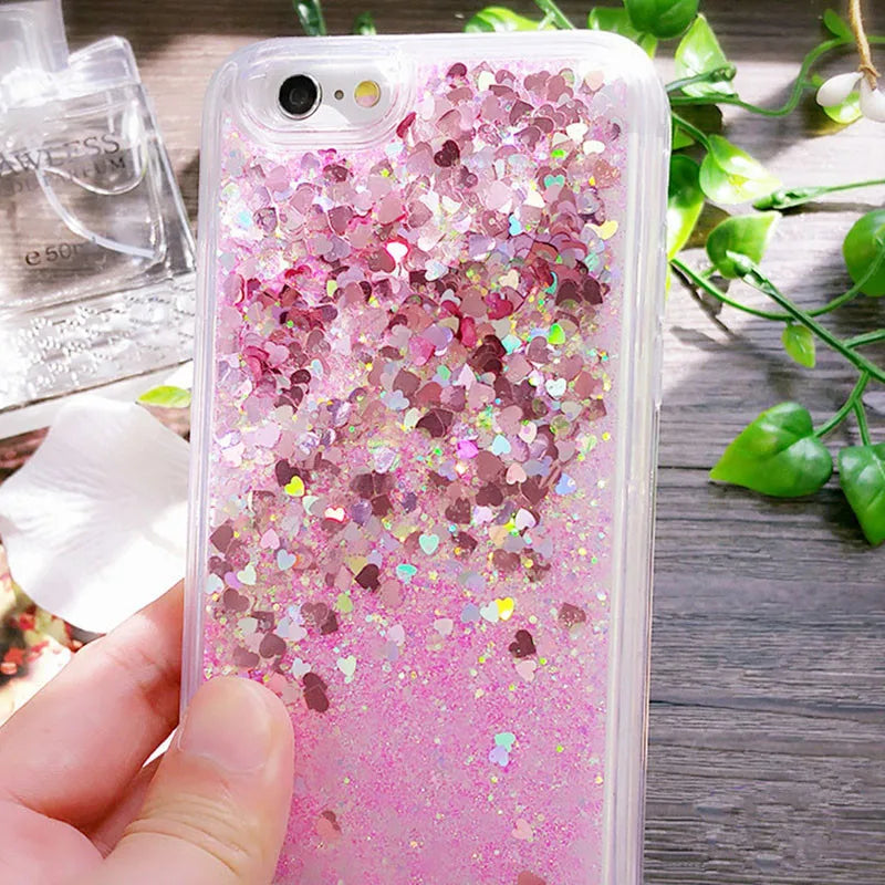 A pink glitter heart star liquid phone case is held by a hand on a wooden surface with green leaves in the background.