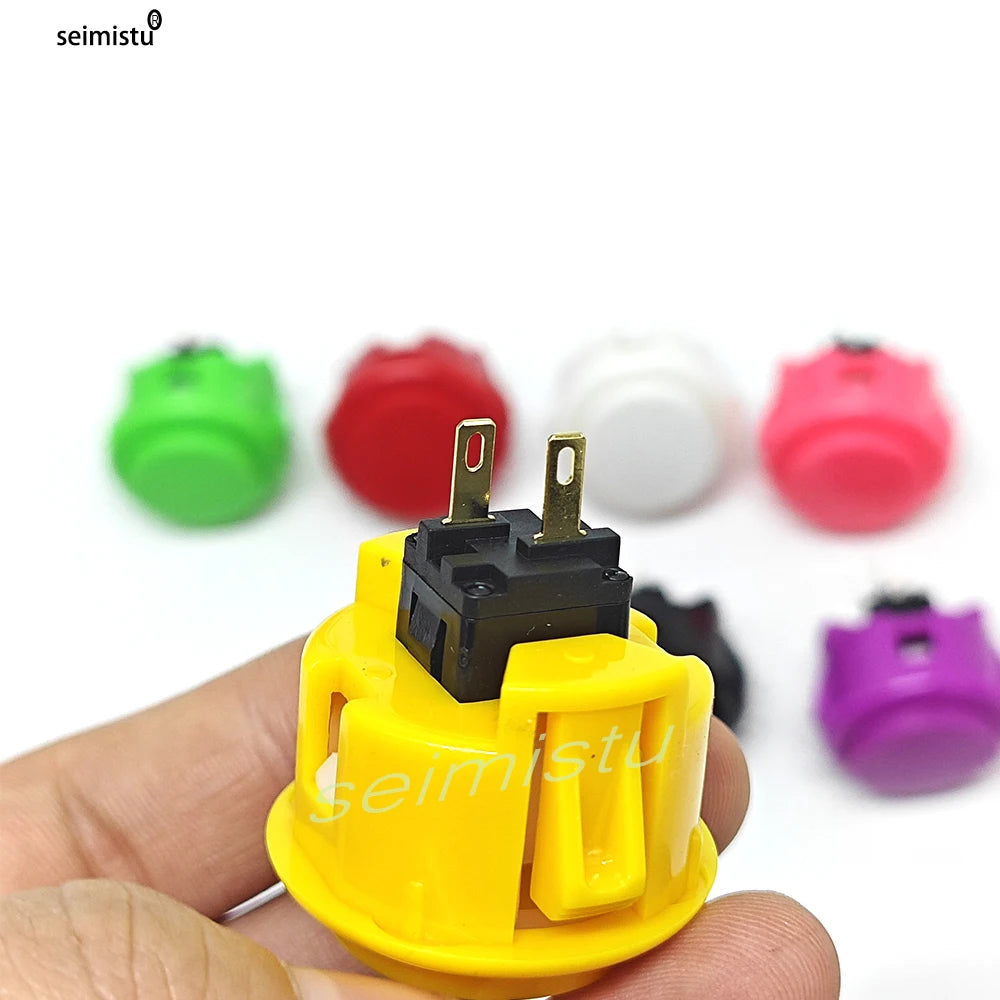 A yellow arcade push button with a black micro switch is held in a hand, surrounded by various colored buttons.