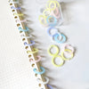 A collection of colorful reusable plastic binding rings in various shades of yellow, pink, and blue are displayed on a white 