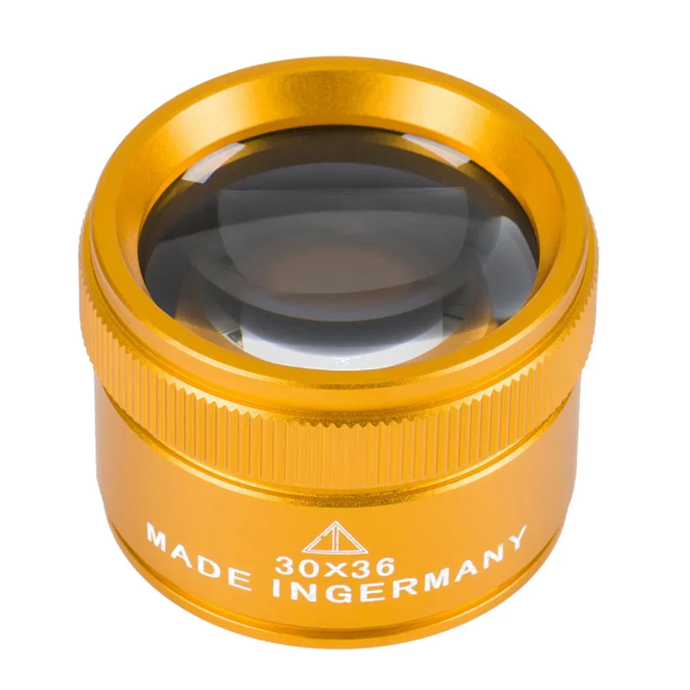 A round, gold-colored magnifying loupe with a glass lens and text indicating it is made in Germany.