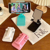 A pink adjustable foldable phone and tablet stand is shown next to a white stand and a blue stand on a wooden table.