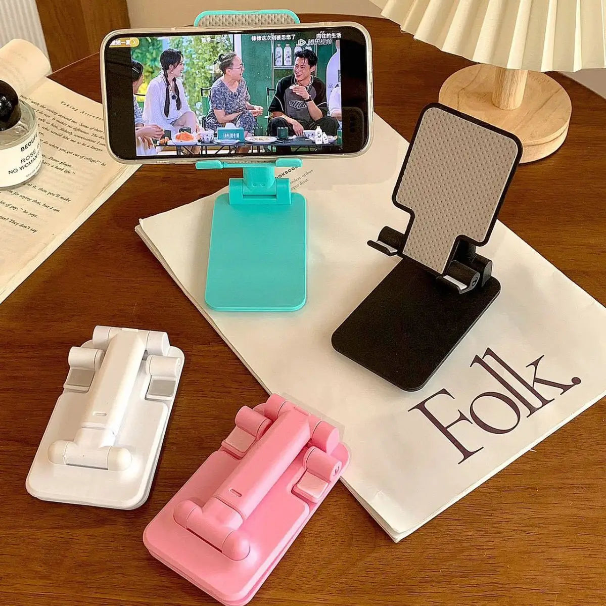 A pink adjustable foldable phone and tablet stand is shown next to a white stand and a blue stand on a wooden table.