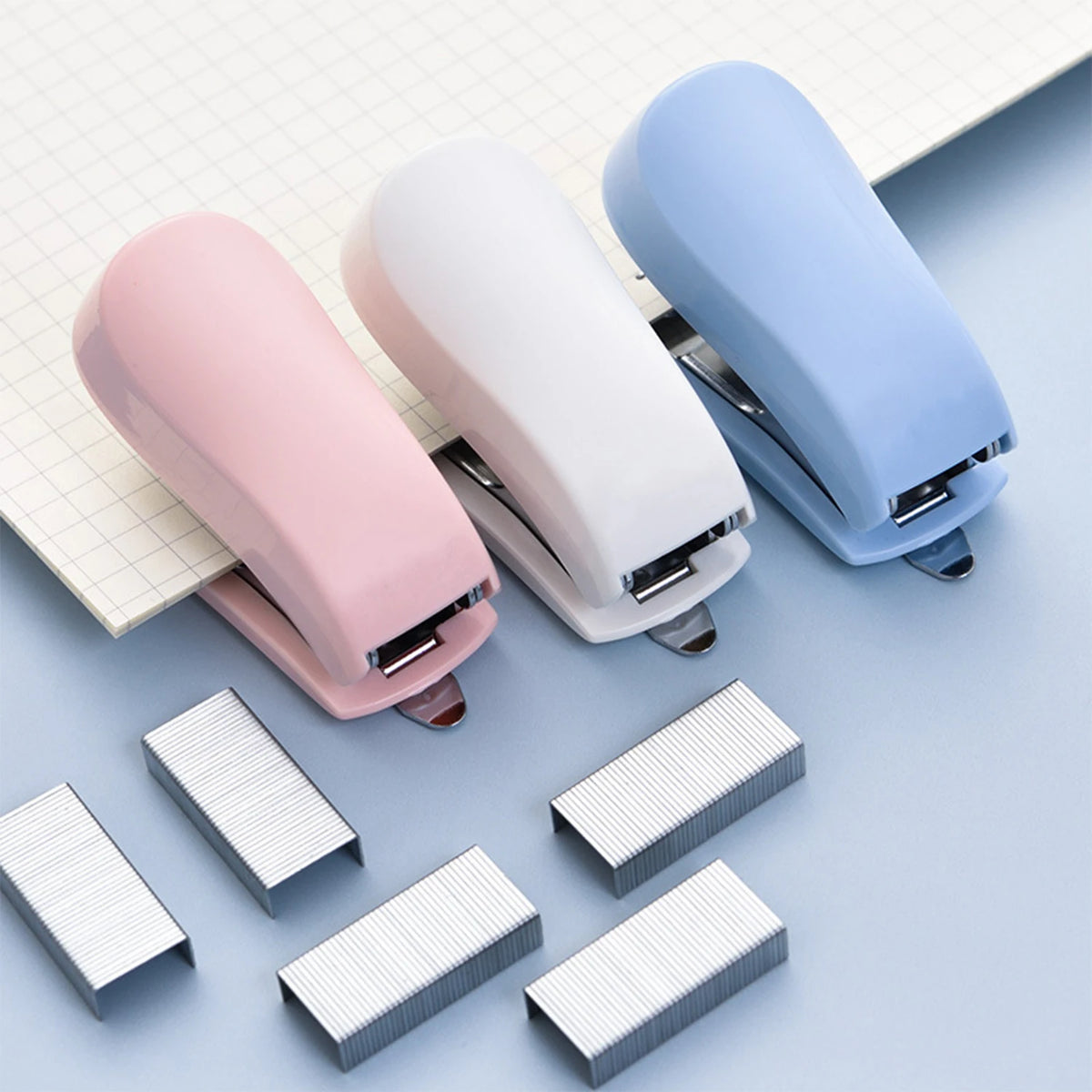 A set of light pink and blue compact mini staplers with staples scattered on a white grid background.