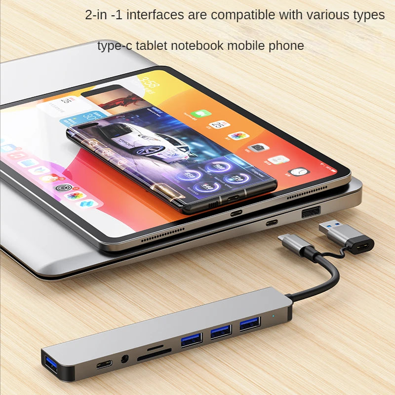 A silver USB-C multiport hub with multiple ports is shown next to a tablet and a smartphone.