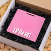 A pink acrylic badge holder with the text CPTURN is placed inside a cardboard box lined with black shredded paper.