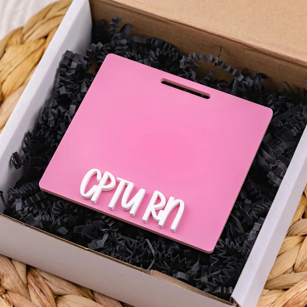 A pink acrylic badge holder with the text CPTURN is placed inside a cardboard box lined with black shredded paper.