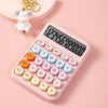 A pink mechanical 12-digit calculator with large round keys is shown on a light pink background next to a small white toy.