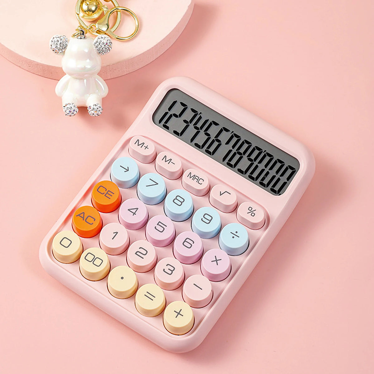 A pink mechanical 12-digit calculator with large round keys is shown on a light pink background next to a small white toy.