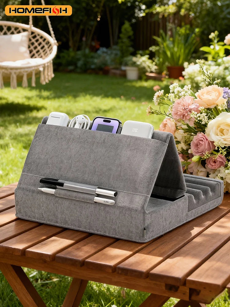 A gray foldable tablet pillow stand with a pocket holding several electronic devices rests on a wooden table outdoors.