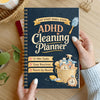 A spiral-bound ADHD cleaning planner with a dark blue cover and gold accents, showing tips for 15-min tasks and room-by-room 