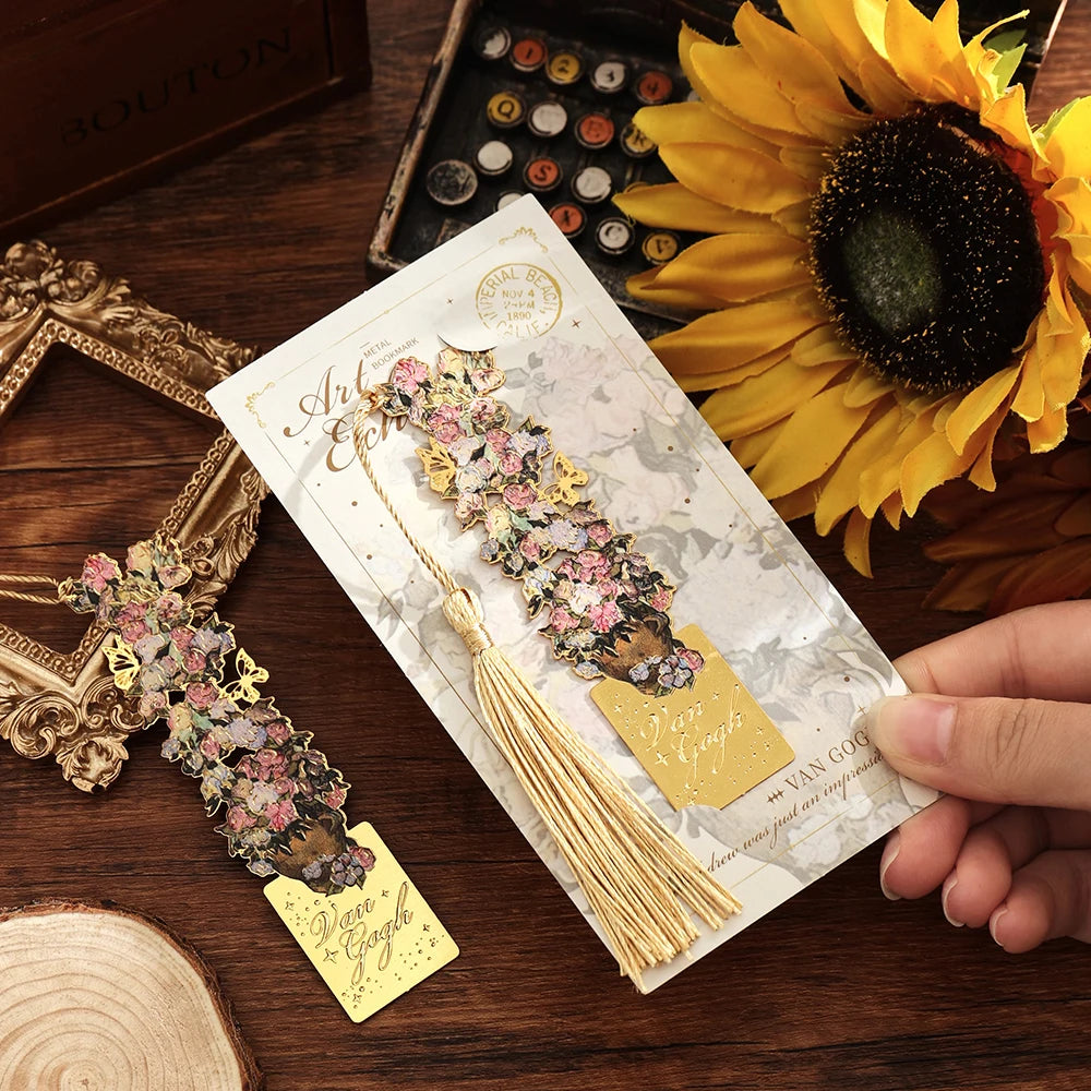A hand holds a vintage artistic metal bookmark with a floral design and gold tassel against a dark wooden background.