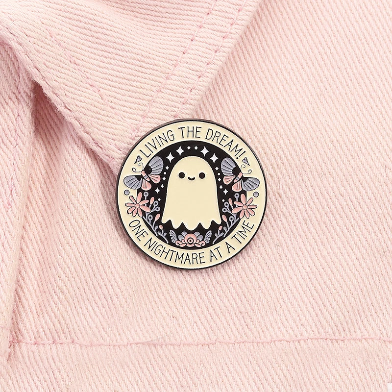 A round enamel pin featuring a white ghost with butterfly wings and text that reads Living the dream! One nightmare at a time
