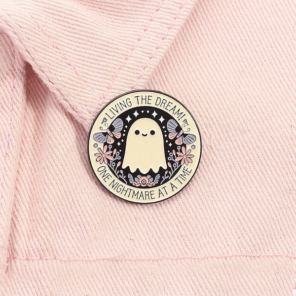 A round enamel pin featuring a white ghost with butterfly wings and text that reads Living the dream! One nightmare at a time