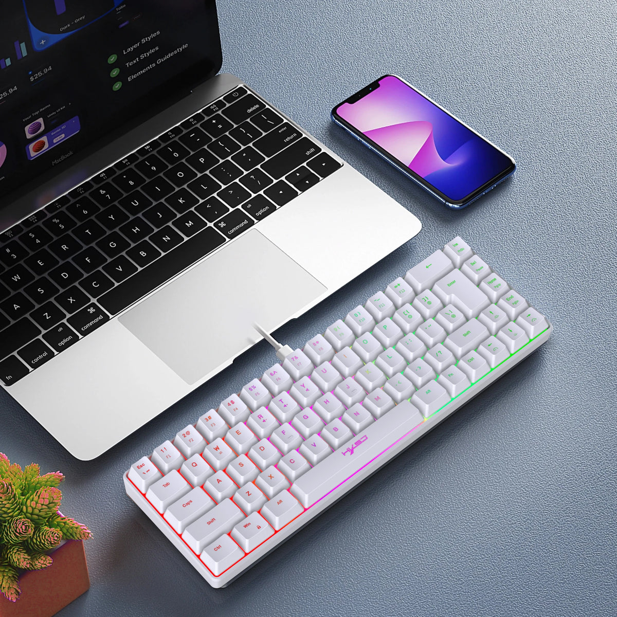 A close-up view of a white 68-key wired RGB gaming keyboard, a smartphone, and a laptop on a gray surface.