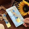 A vintage artistic metal bookmark with a tassel is displayed next to a sunflower on a wooden surface.