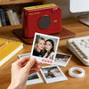 A hand holds a red smart Bluetooth label maker device next to various photo prints and paper on a wooden table.