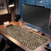 A leopard print XXL gaming mouse pad is displayed on a wooden desk next to a monitor and speakers.