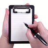 A hand holds a black pocket-sized clipboard with a secure clip, writing on the lined paper with a black pen.
