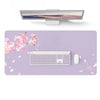 A silver laptop is displayed above a large purple desk mat featuring pink blossoms and a white keyboard and mouse.