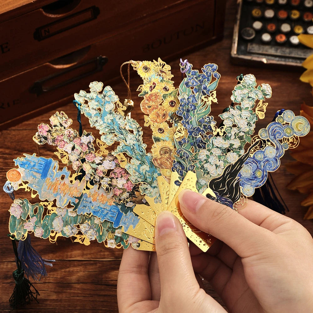 A hand holds a vintage artistic metal bookmark with a gold leaf design and blue floral elements.