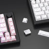 A close-up view of a black mechanical keyboard and various white and pink PBT keycaps and legends scattered on a dark surface